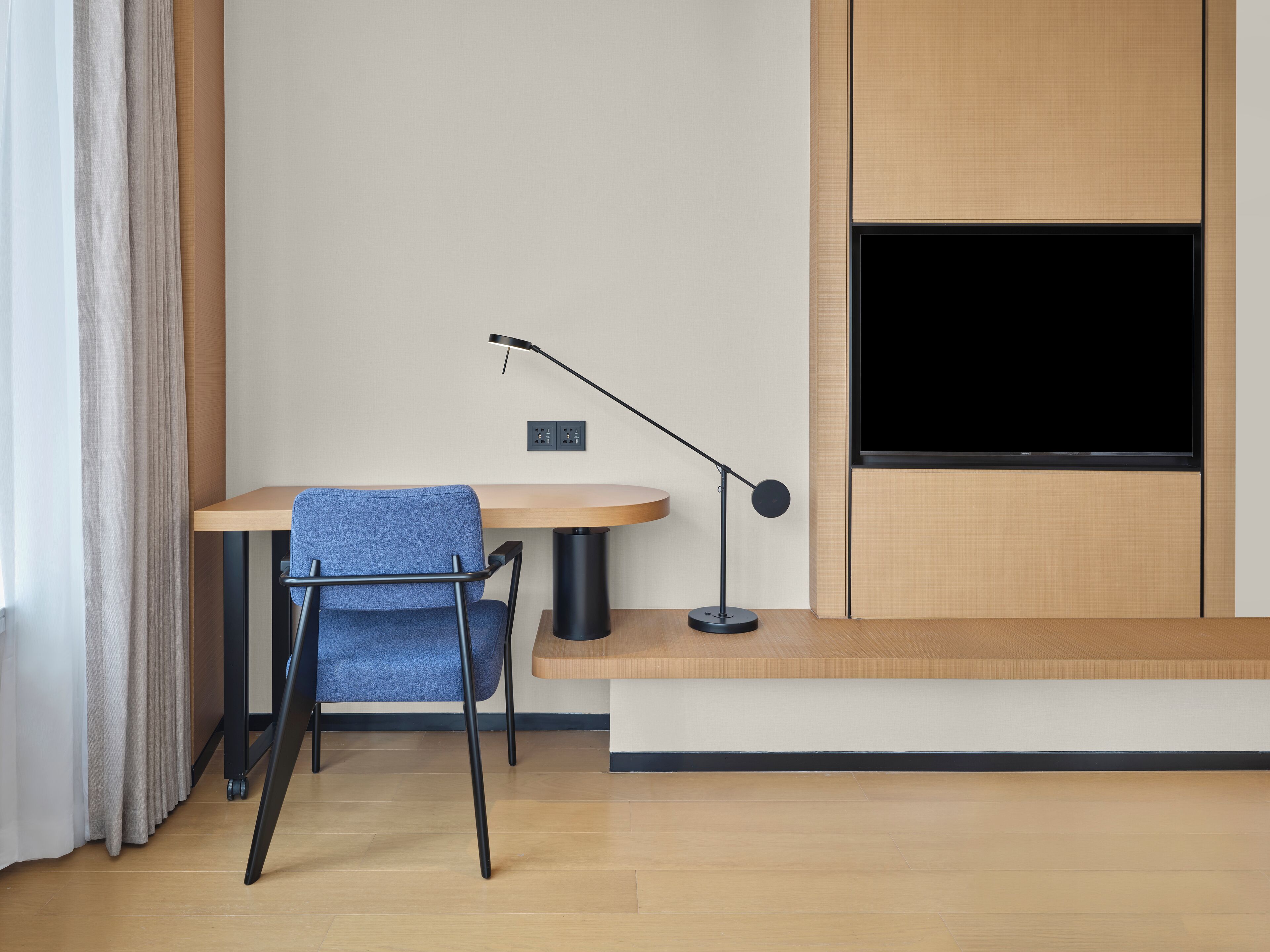 Une chambre moderne avec un bureau en bois, une chaise bleue, un lampadaire et un téléviseur fixé au mur.