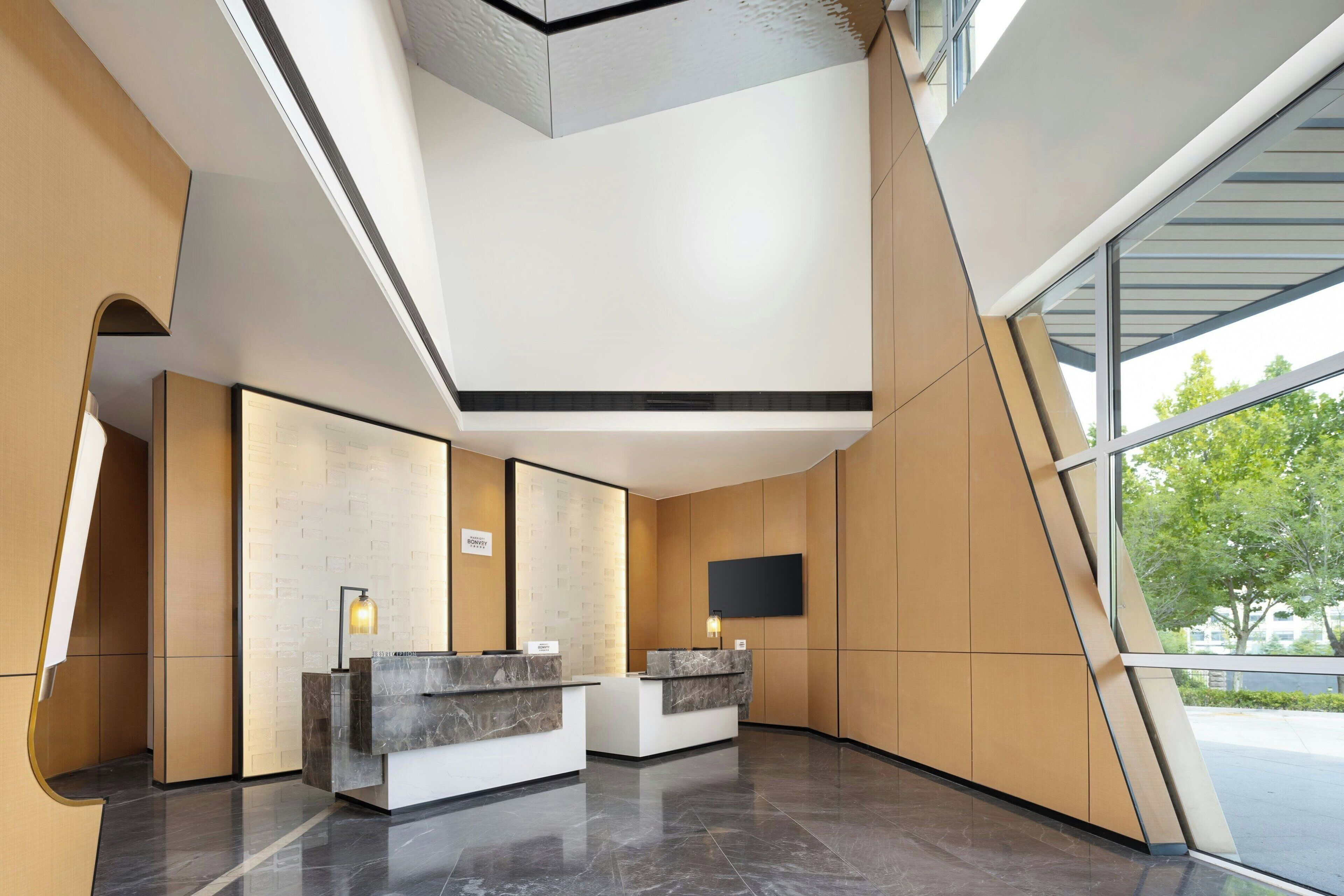 A modern reception area with two marble reception desks, a flat-screen TV mounted on the wall, and a large window providing a view of the outdoors.