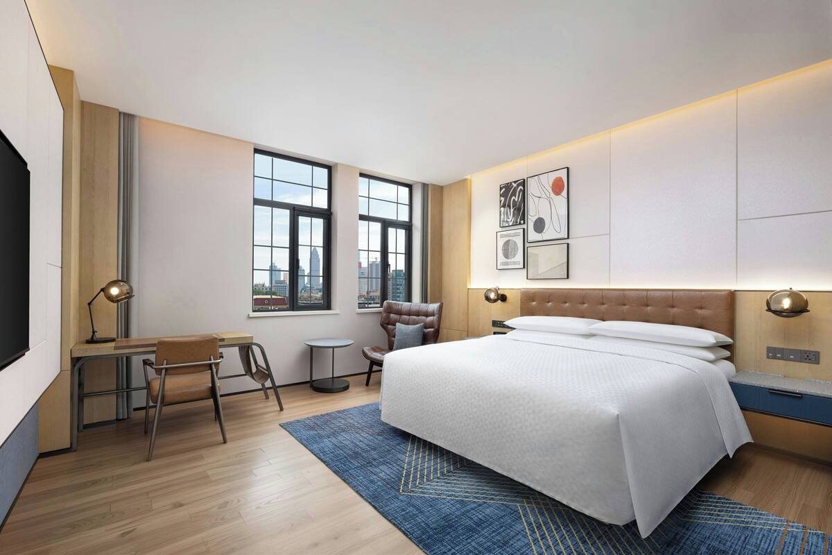 A modern hotel room with a large bed, a desk, and a chair. There are windows with a city view, a wall-mounted artwork, and a small round table.