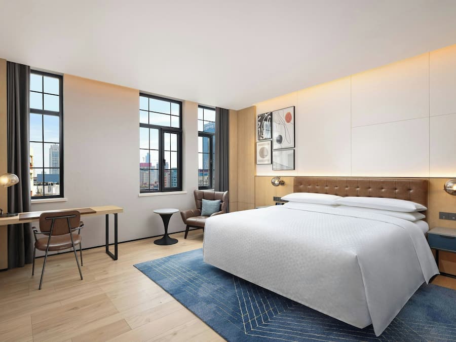 A modern hotel room with a large bed, a desk, a chair, and a view of the city through large windows.