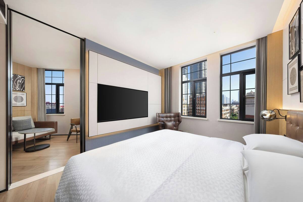 A modern hotel room with a large bed, a flat-screen TV mounted on the wall, a seating area with a sofa and armchair, and large windows offering city views.