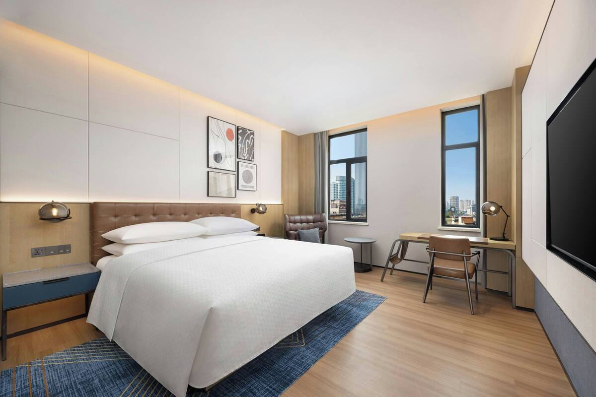 A hotel room with a large bed, a desk, a chair, and a TV. There are windows and artwork on the walls.