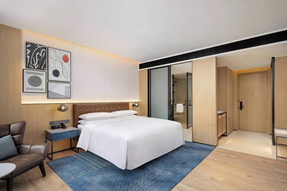 A modern hotel room with a large bed, a bedside table with a clock, a chair, and a blue rug. There is a wardrobe and a sliding door closet.
