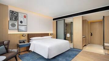 A modern hotel room with a large bed, a bedside table with a clock, a chair, and a blue rug. There is a wardrobe and a sliding door closet.