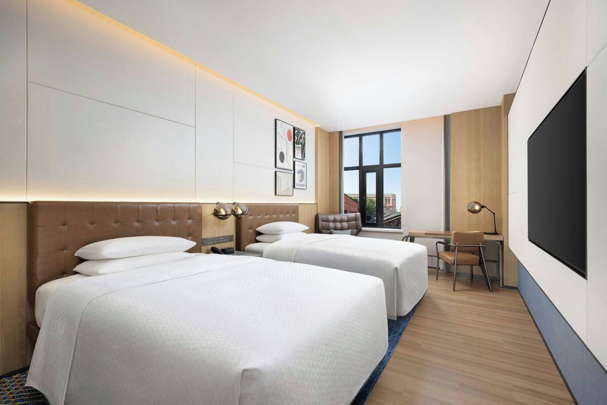 A hotel room with two beds, a desk, a chair, a TV, and a window with a view of buildings.