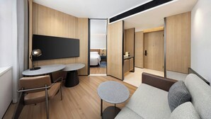 Ett modernt hotellrum med en soffa, två små bord, en TV och ett separat sovrum med en säng och ett skrivbord.