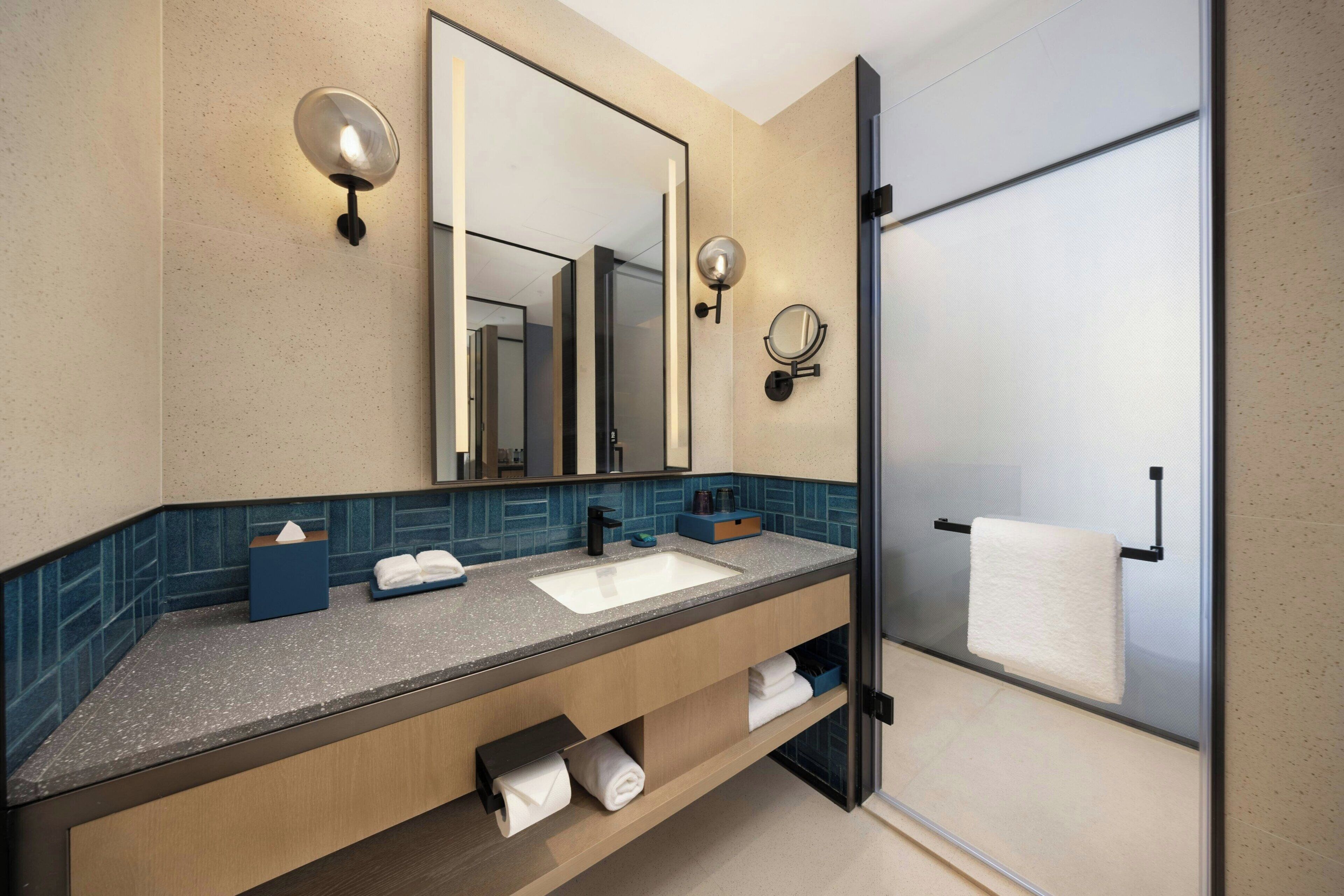 Une salle de bain moderne avec un grand miroir, deux appliques murales, un lavabo et une cabine de douche en verre.