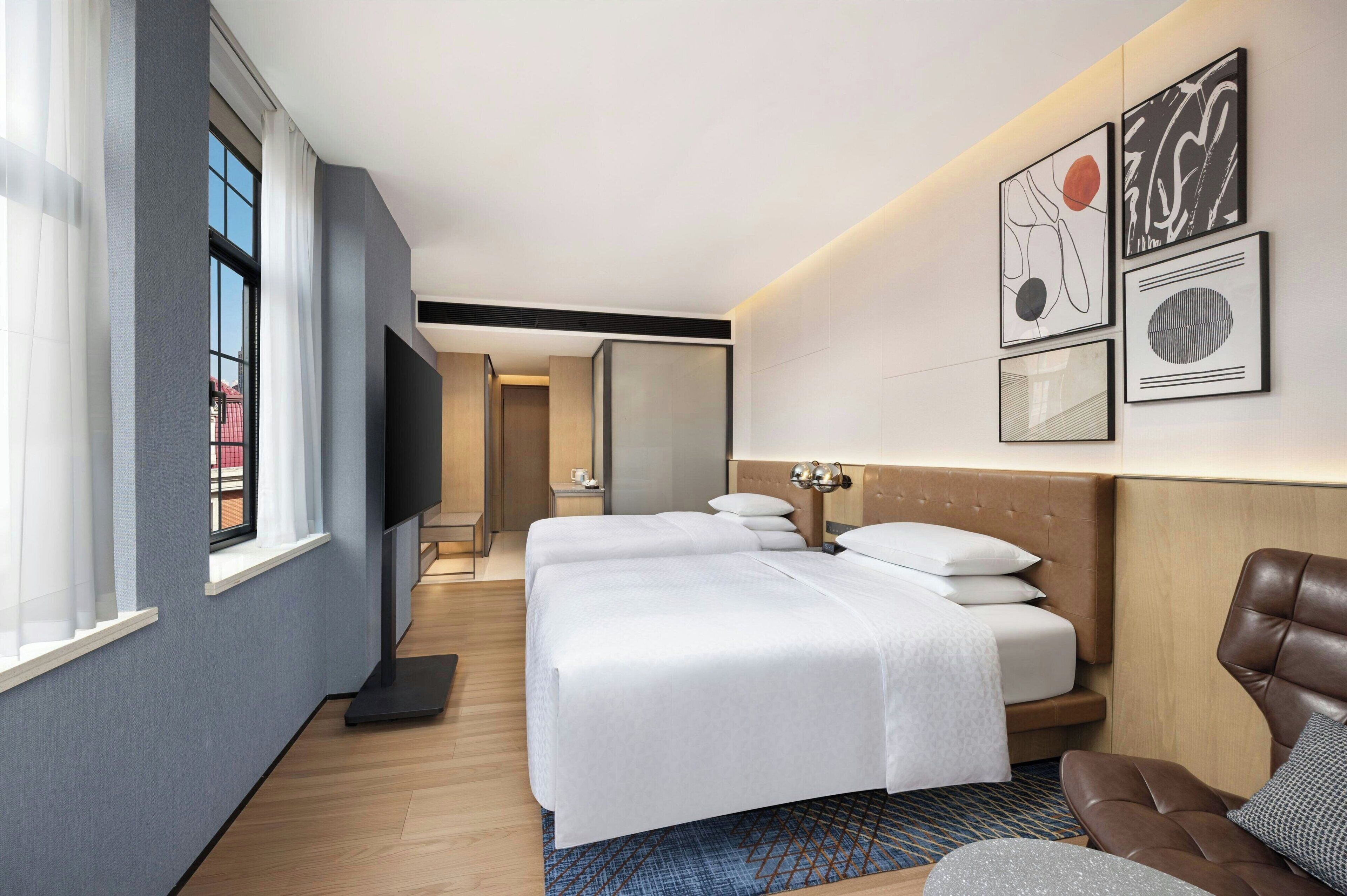 Quarto de hotel com duas camas, uma poltrona de couro marrom e obras de arte na parede.