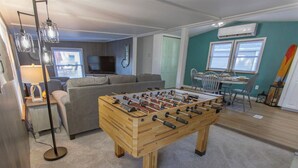 A room with a wooden foosball table, a grey sofa, a TV, and a dining area with chairs and a table. - Cozy 1-bedroom mobile home on the beautiful Crooked lake (Angola)