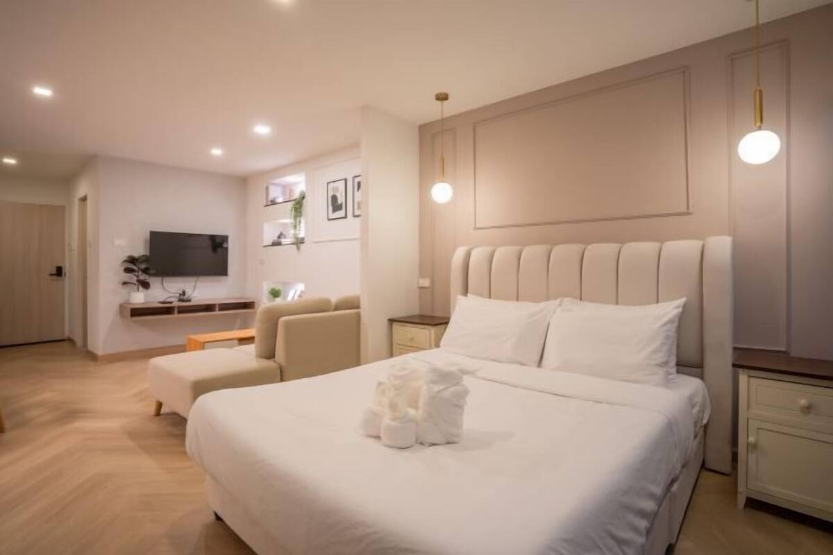 A modern hotel room with a large bed, a bedside table, a TV mounted on the wall, and a seating area with a sofa and chairs.