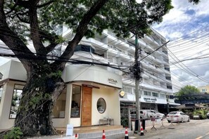 Un hotel con fachada moderna, un gran árbol en frente y un estacionamiento con coches.