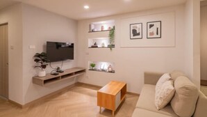 A modern living room with a flat-screen TV, wooden coffee table, beige sofa, and wall-mounted shelves with decorative items.