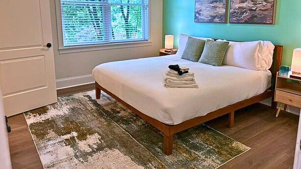 A bedroom with a bed, a window with blinds, a bedside table with a lamp, and two wall-mounted paintings. - Well-Equipped 2BR - Greenbay 301 (Evanston)