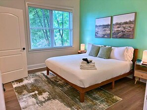 Un dormitorio con cama, ventana con persianas, mesita de noche con lámpara y dos cuadros colgados en la pared.