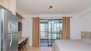 A modern hotel room with a bed, a desk, and a view of a cityscape through a balcony.