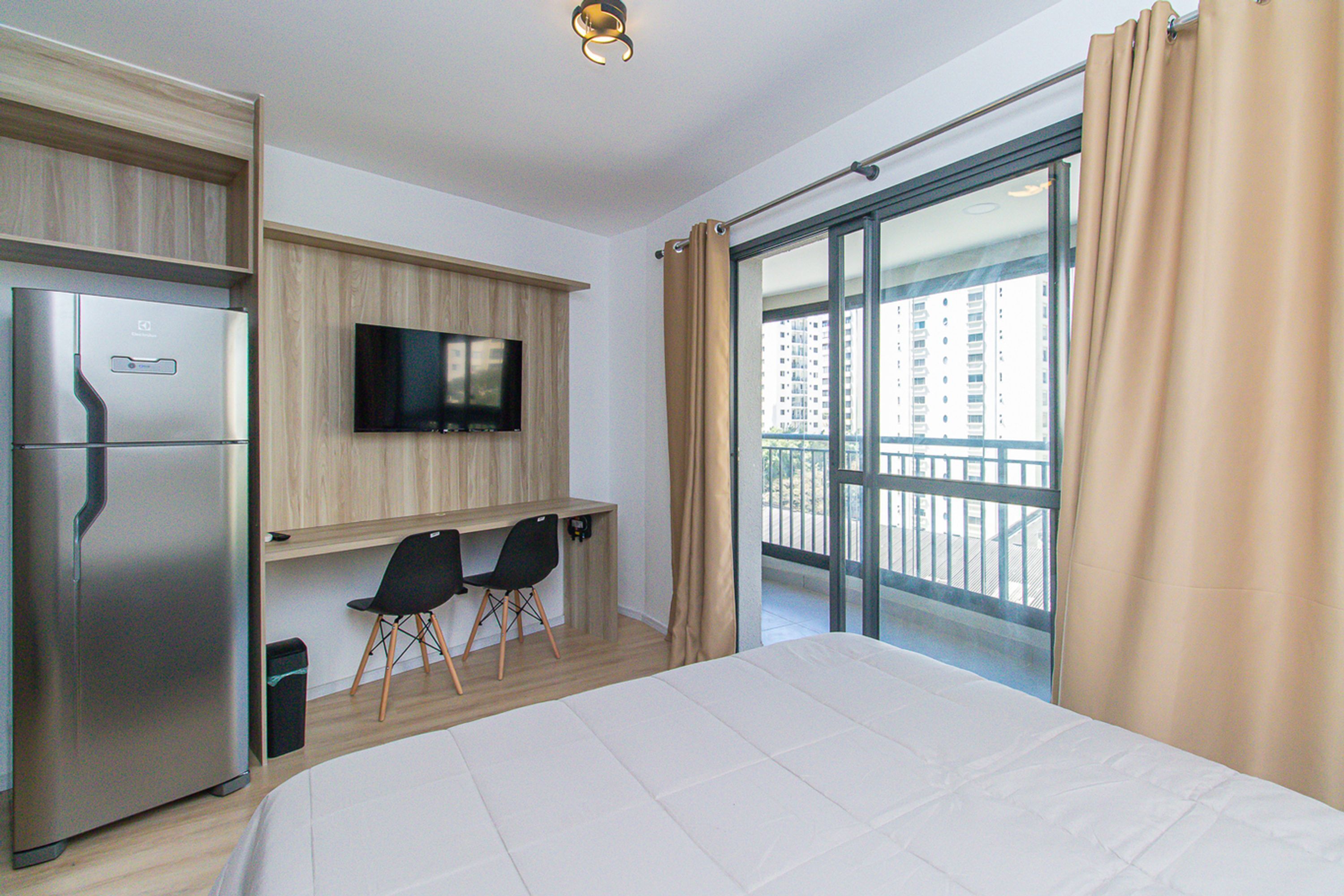 A hotel room with a bed, a desk with two chairs, a television, and a balcony with curtains.