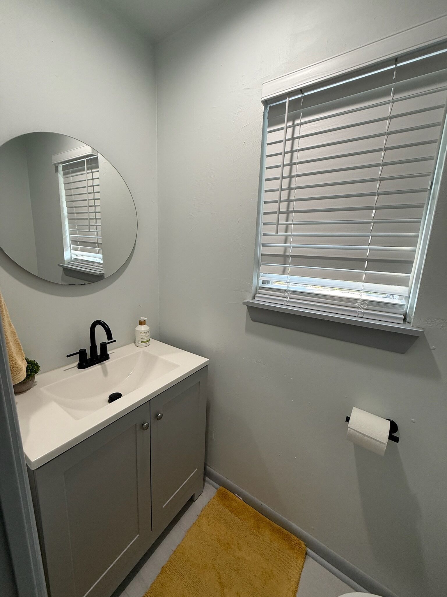 A bathroom with a white sink, a mirror, a toilet, and a window with blinds.