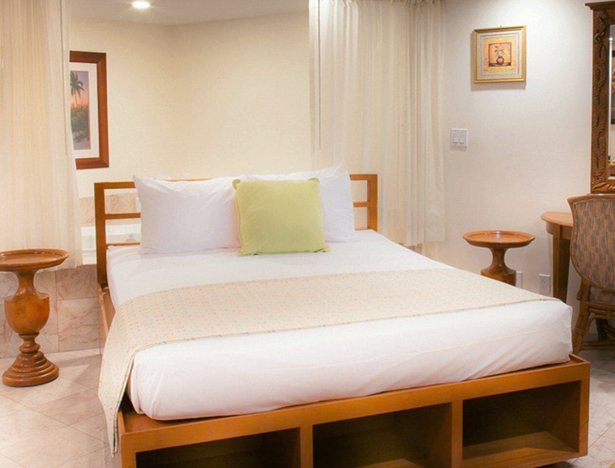 A neatly made bed with a wooden frame, a green pillow, and a white bedspread. There are two small wooden side tables, one with a round table on it.
