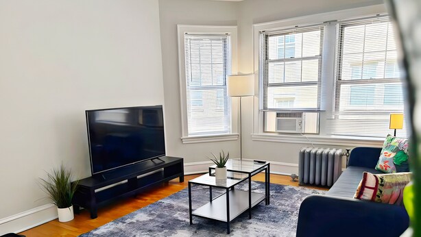 A modern living room with a flat-screen TV, a glass coffee table, a sofa, and a radiator. - Perfectly Located 1BR - Hinman D2 (Evanston)