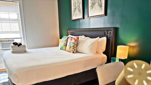 A neatly made bed with white linens and colorful pillows, a bedside table with a lamp, and framed artwork on the wall. - Perfectly Located 1BR - Hinman D2 (Evanston)