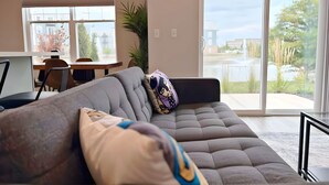 Un salon moderne avec un canapé gris, un coin repas comprenant une table et des chaises, et une grande porte-fenêtre coulissante donnant sur un balcon avec vue sur l’extérieur.