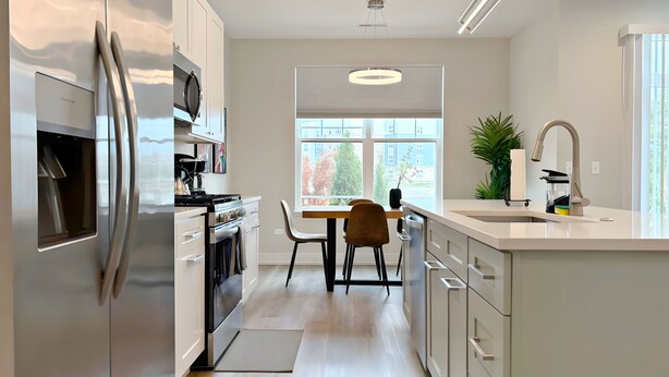 Une cuisine moderne avec des meubles blancs, des appareils en acier inoxydable, un coin repas avec une table et des chaises, et une grande fenêtre offrant une vue sur l’extérieur.