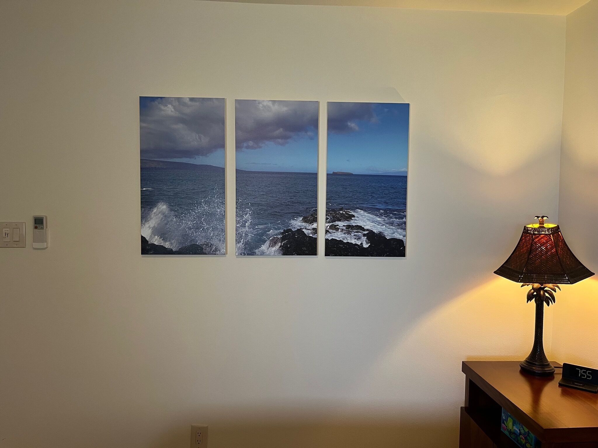 A wall with three framed photographs of the ocean, a wall socket, and a lamp on a nightstand.