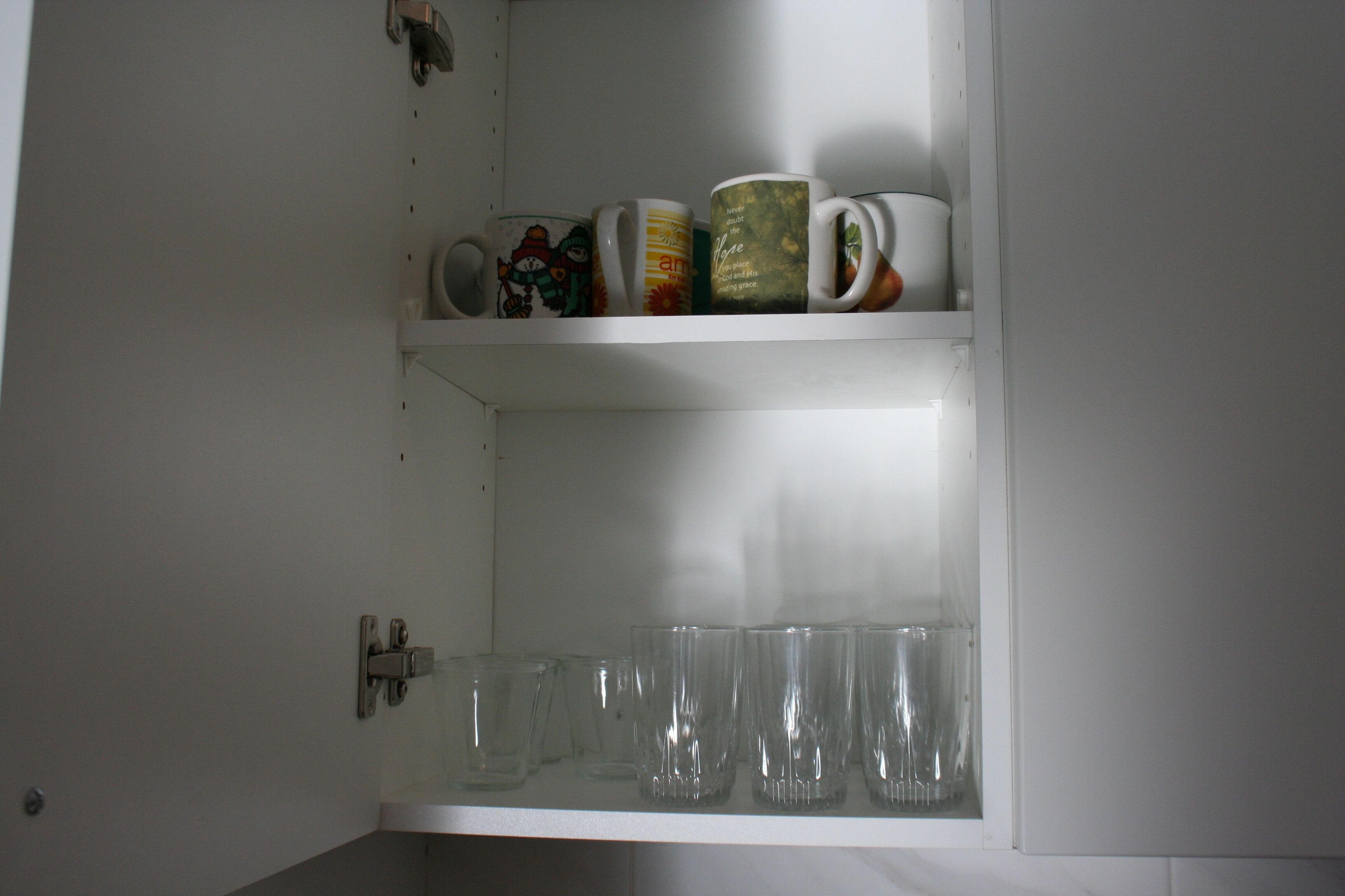 Un armario de cocina con dos estantes; el estante superior contiene tazas decorativas y el inferior, vasos de vidrio.