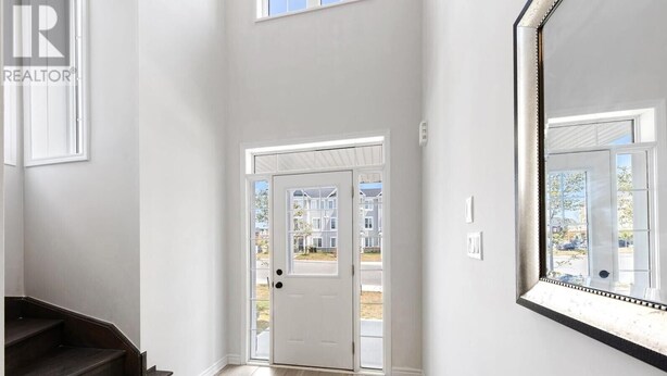 A clean, modern hallway with a white door leading outside, a large mirror on the right, and a dark staircase on the left.