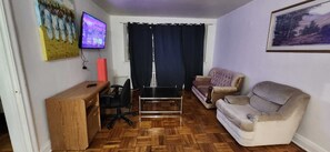 Ein Wohnzimmer mit einem Holztisch, einem schwarzen Bürostuhl, einem Couchtisch mit Glasplatte, einem beigen Sofa und einem an der Wand montierten Fernseher.