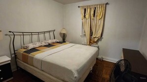 Ein Einzelbett mit Kopfteil, ein Nachttisch mit Lampe, ein Heizkörper und ein Fenster mit Vorhängen.
