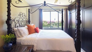 A bedroom with a large bed, dark wooden headboard, a ceiling fan, and a view of flowers outside.