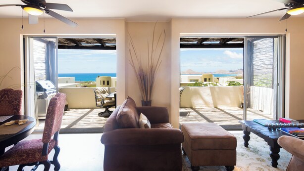 A living room with a sofa, armchair, and ottoman, a coffee table, and a ceiling fan. There is a view of the ocean through large glass doors.