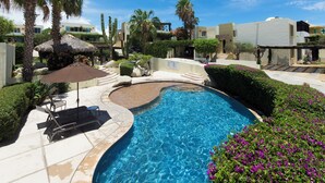 A swimming pool with a curved design, surrounded by palm trees, a thatched-roof structure, and a flower garden.
