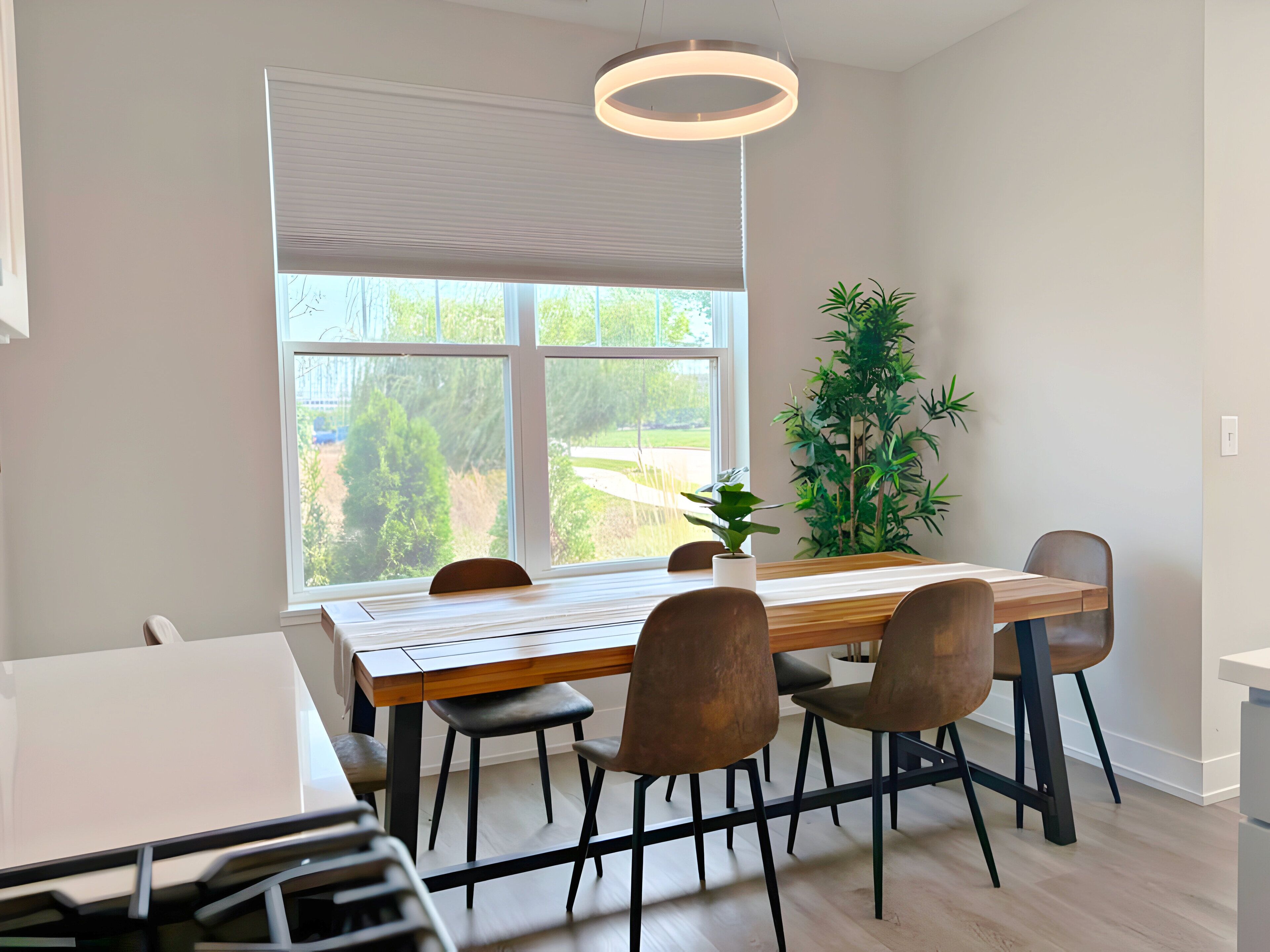 A modern dining area with a wooden table and chairs, a large window with blinds, a potted plant, and a hanging light fixture.