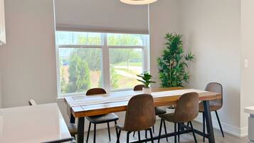 A modern dining area with a wooden table and chairs, a large window with blinds, a potted plant, and a hanging light fixture.