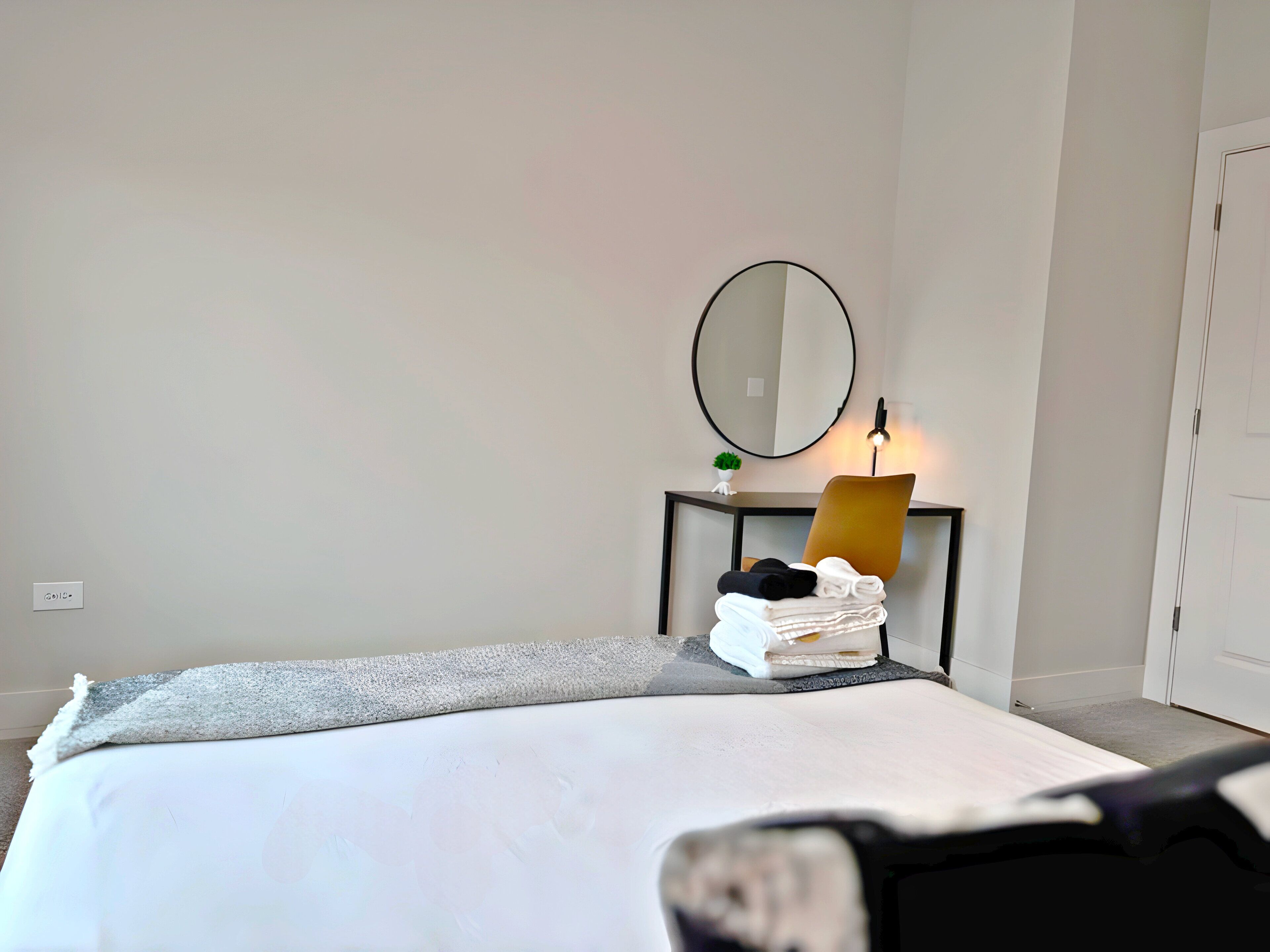 A neatly made bed with a white comforter and a gray throw blanket. There is a small table with a mirror, a lamp, and a folded towel.
