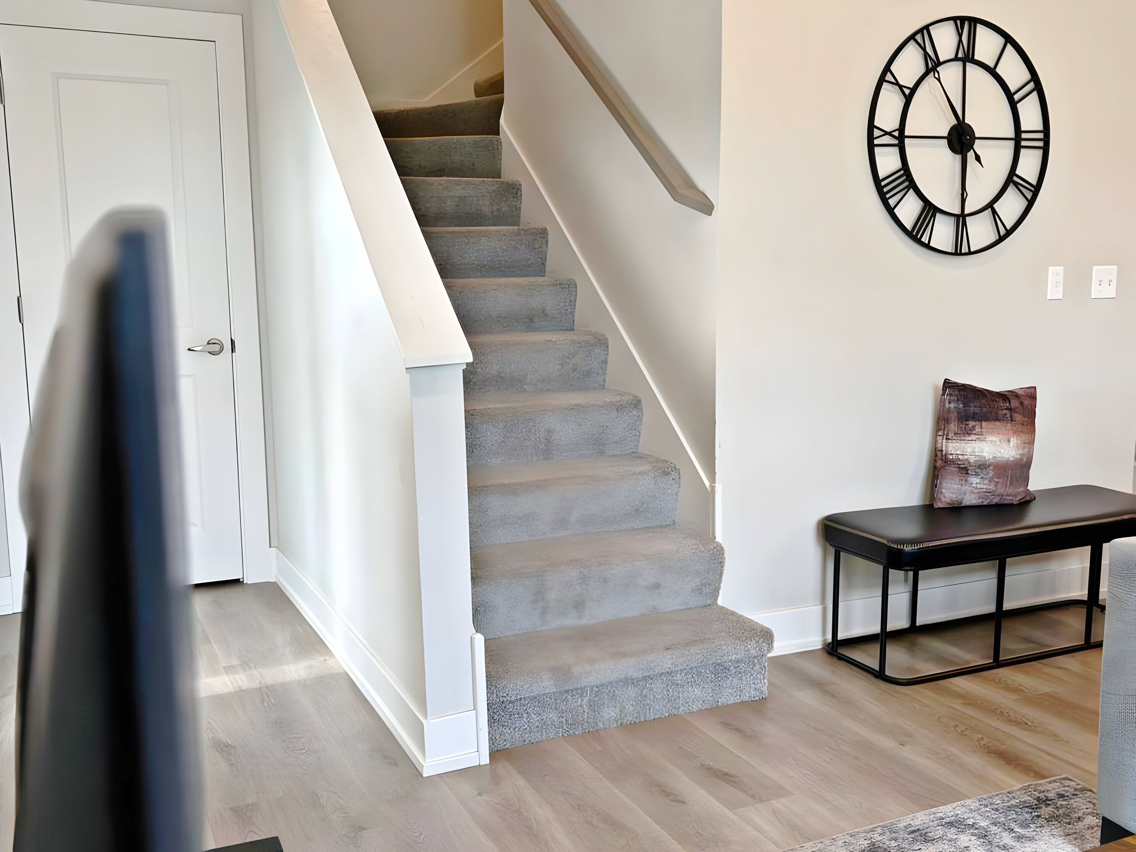 A staircase with a handrail, a wall-mounted clock, a small table with a cushion, and a door.