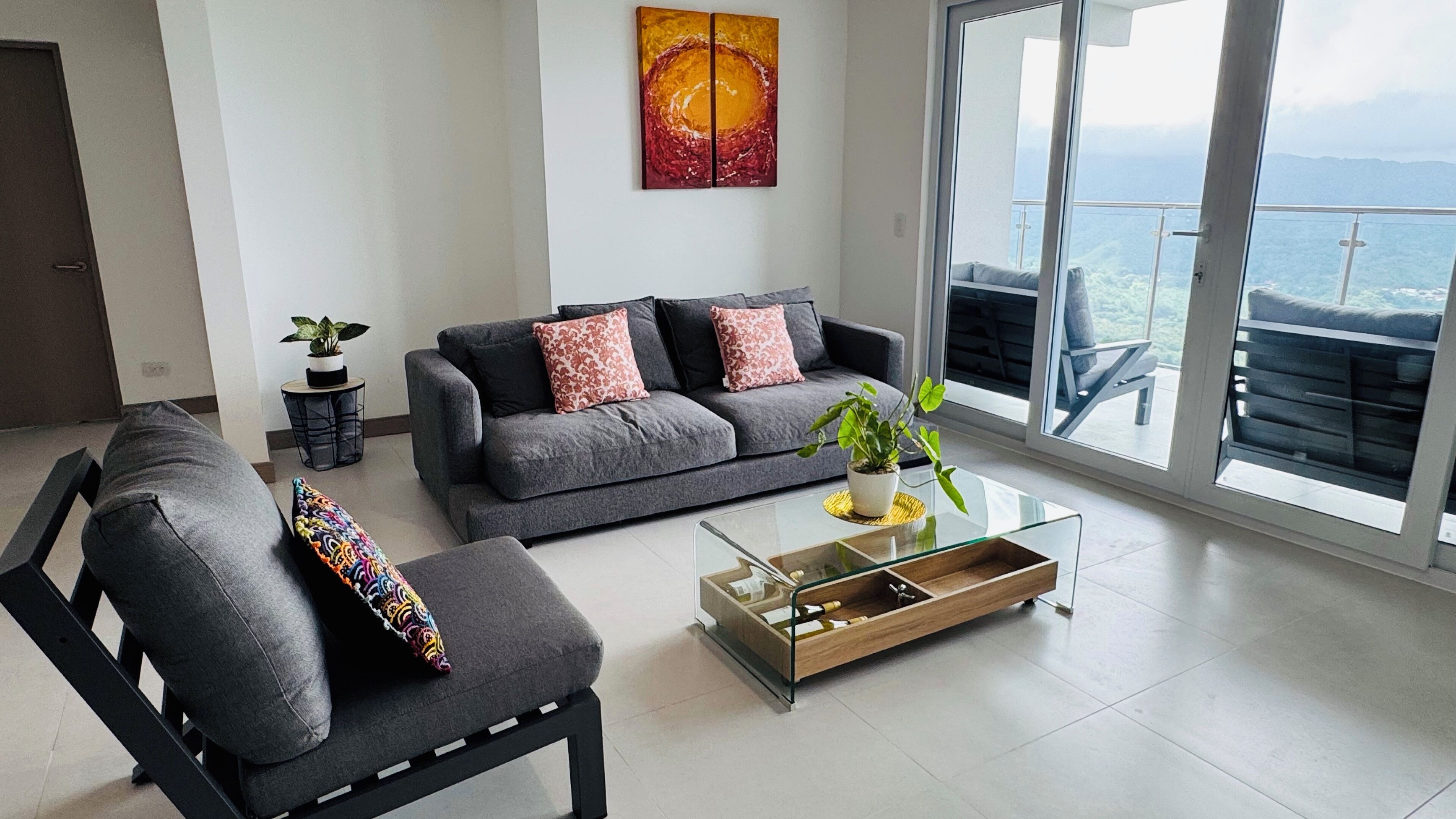 Uma sala de estar moderna com um sofá cinzento, uma poltrona preta e uma mesa de centro de vidro. Há uma grande janela com vista para o mar e uma varanda.