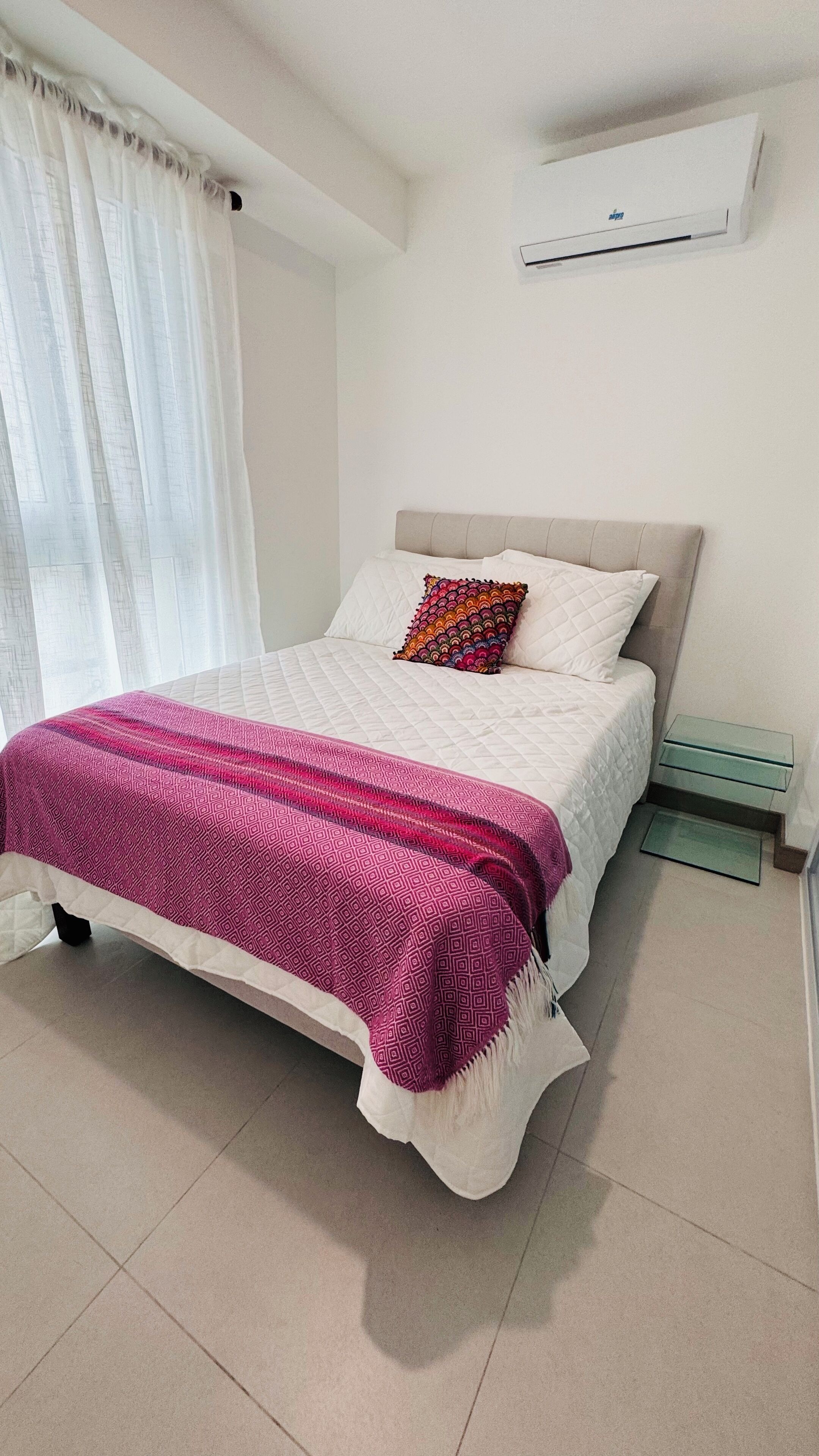 A bedroom with a bed, a bedside table, a window with curtains, and an air conditioning unit on the wall.