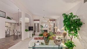 A spacious living room with a glass coffee table, a dining area with chairs, a kitchen with white cabinets, and a large potted plant.