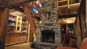Un intérieur chaleureux avec une cheminée en pierre, des étagères en bois et un plafond haut avec des poutres apparentes.