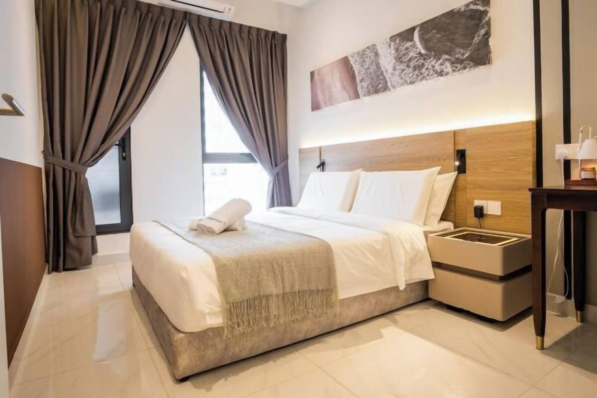 A hotel room with a bed, a nightstand, a wall-mounted lamp, and a window with curtains.