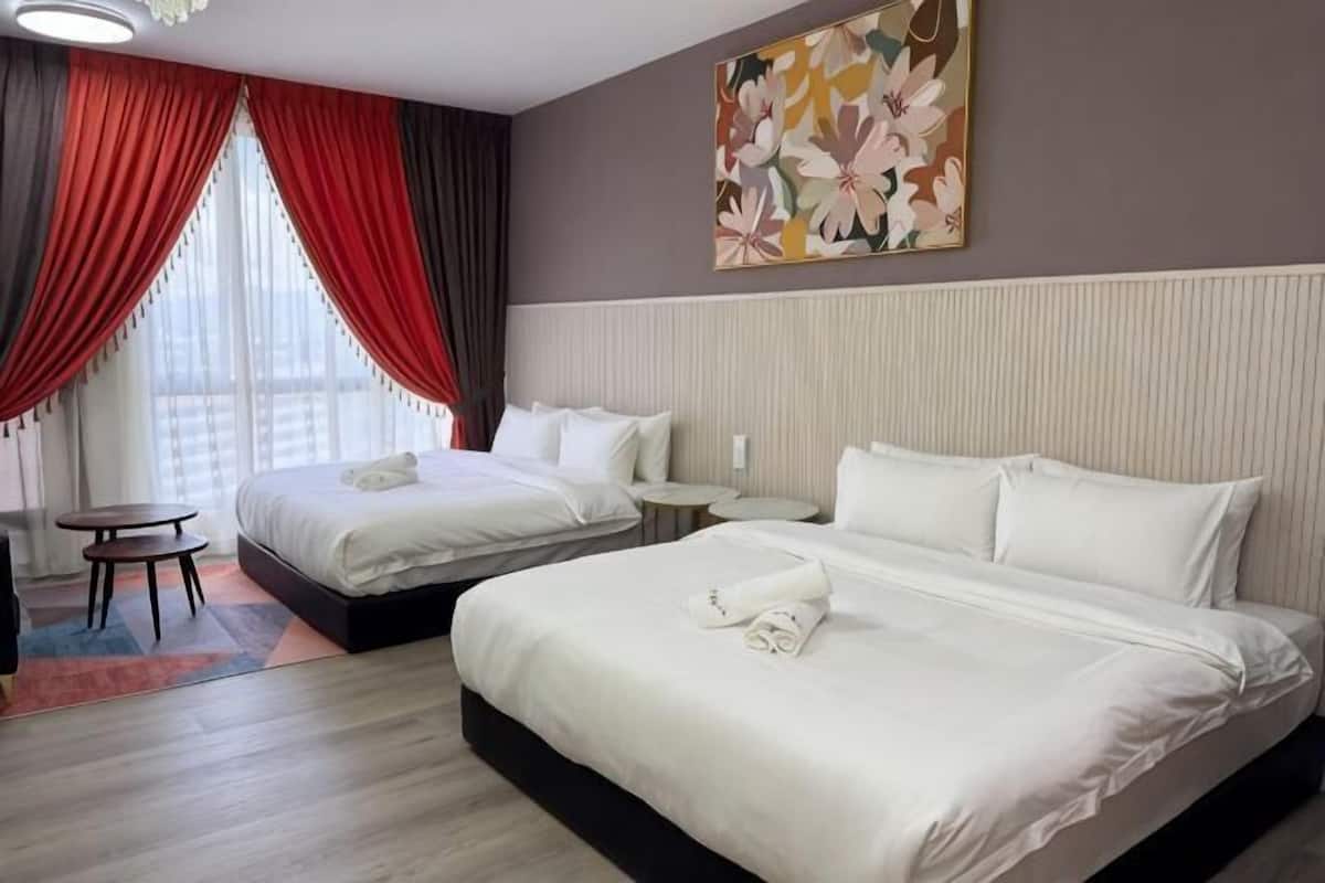 A hotel room with two beds, a small table, a chair, and a window with red curtains.