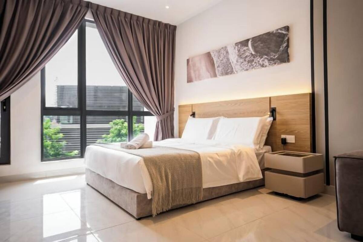A modern bedroom with a large bed, a nightstand, and a view of greenery through large windows.