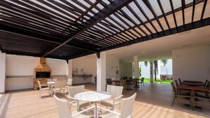Une terrasse extérieure dotée d’une cheminée en briques, de meubles en osier blancs et d’un espace abrité avec un toit noir.