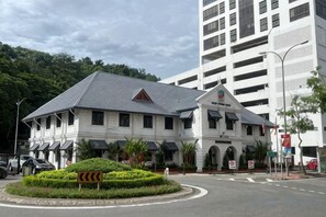 A building with a central entrance, multiple windows, and a landscaped area with a sign indicating a curve ahead. - Jesselton Quay - Sunset Ocean View Apartments by Quality Home Living (Kota Kinabalu)