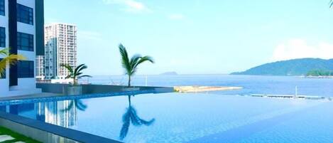 An infinity pool with a view of the sea and a distant island, surrounded by modern buildings.