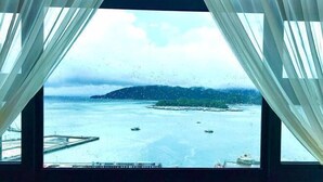 A view through a window with white curtains, featuring a body of water, boats, and a distant island.