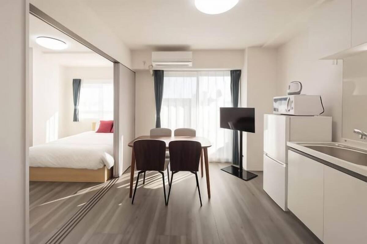 Une chambre d’hôtel de taille réduite comprenant un lit, une petite table à manger avec des chaises, un téléviseur, un four à micro-ondes et une kitchenette.